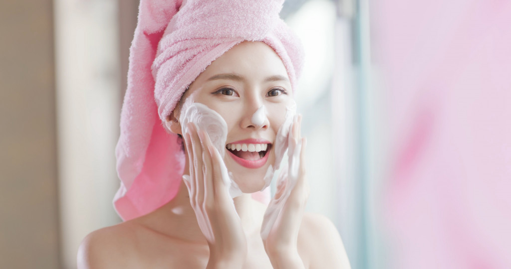 Young woman using a skin cleanser while wearing a towel on her head.
